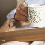 Häusliche Pflege mit Heilpflanzen und Hausmitteln: Fotoausschnitt eines Pflegebedürftigen, der im Bett ein Tablett mit einer großen Tasse Kräutertee hält.